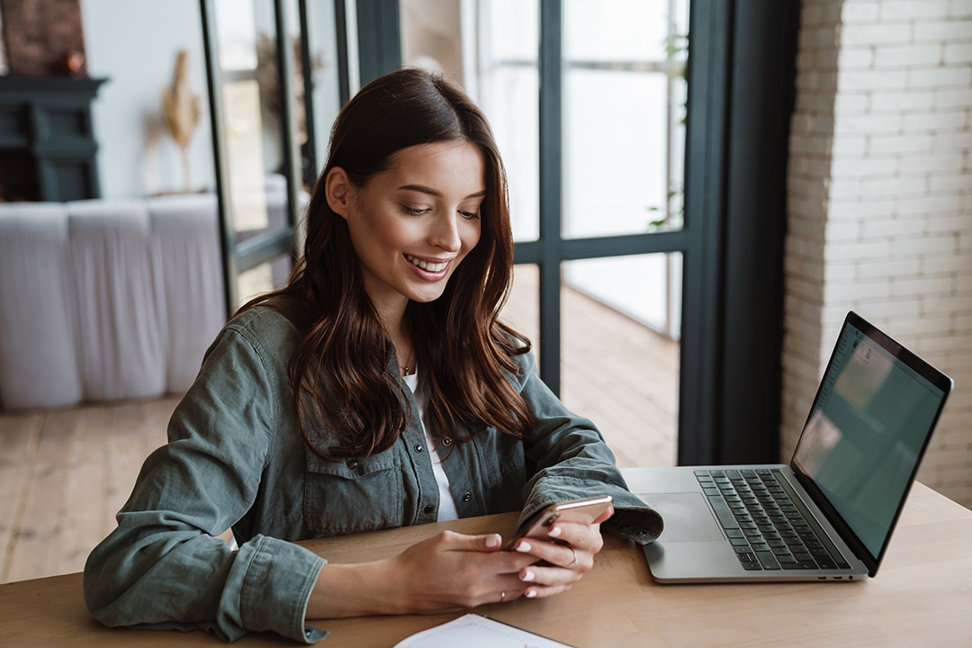 Beautiful smiling woman using cellphone while working with laptop indoors